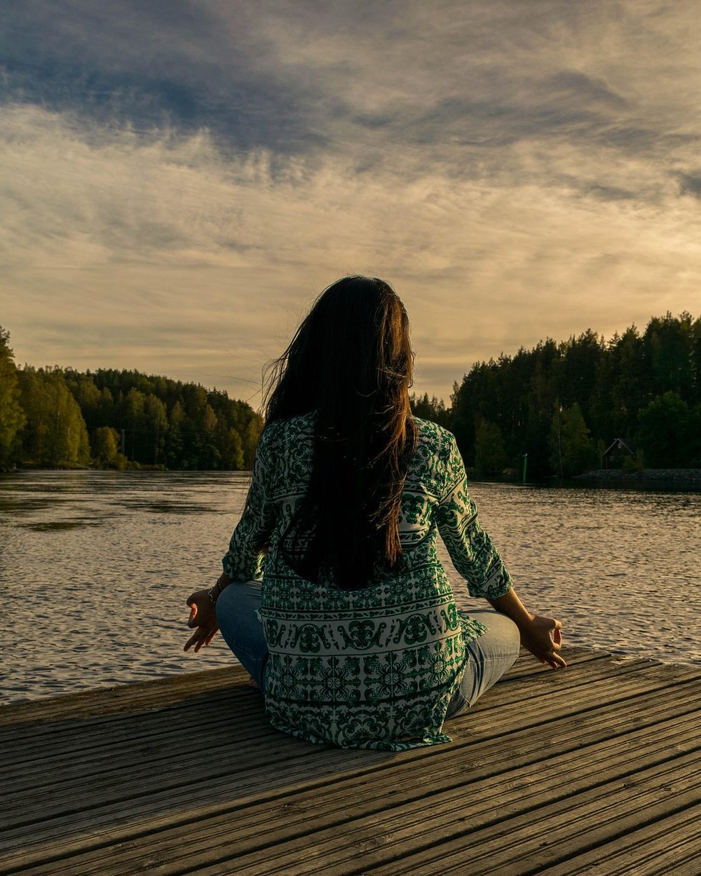 A woman sits cross-legged in meditation at the end of a wooden dock, looking out over a lake at dusk