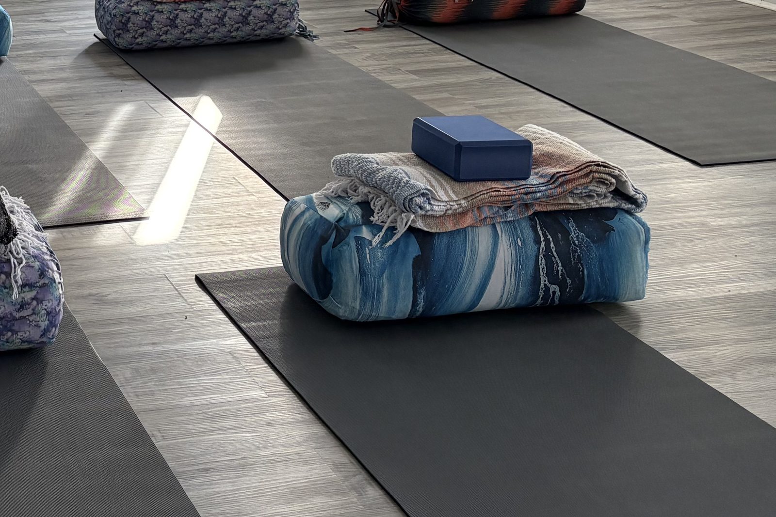A yoga bolster topped with folded blankets and a blue block, set on a mat in a sunlit room