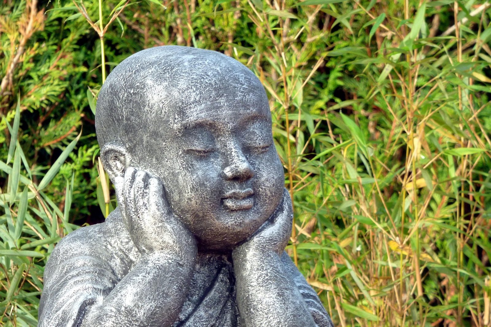 A stone statue of a smiling monk resting his cheeks in his hands, eyes closed, against tall grass