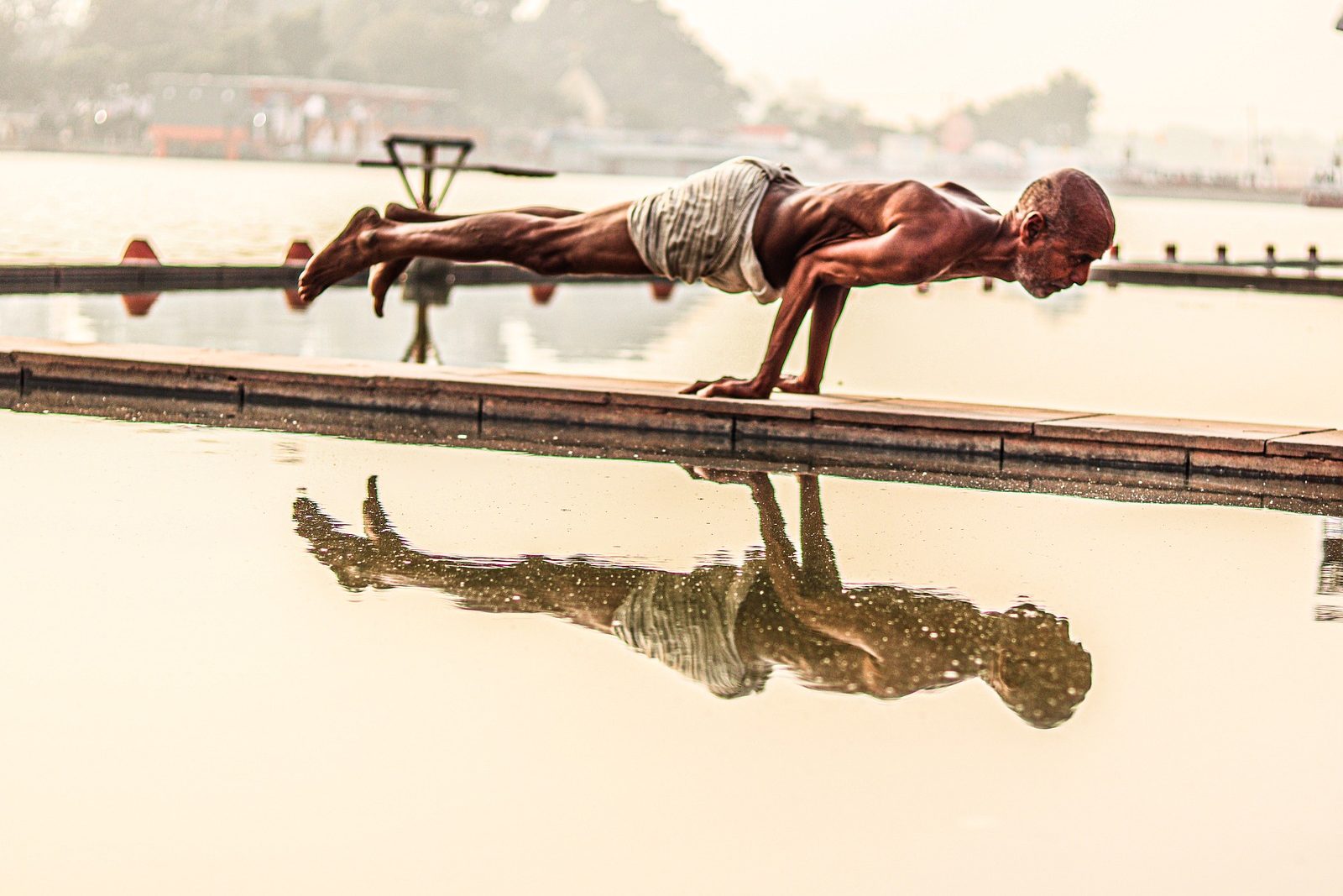 A man holds a plank pose on a narrow walkway over still water, mirrored in the reflection below